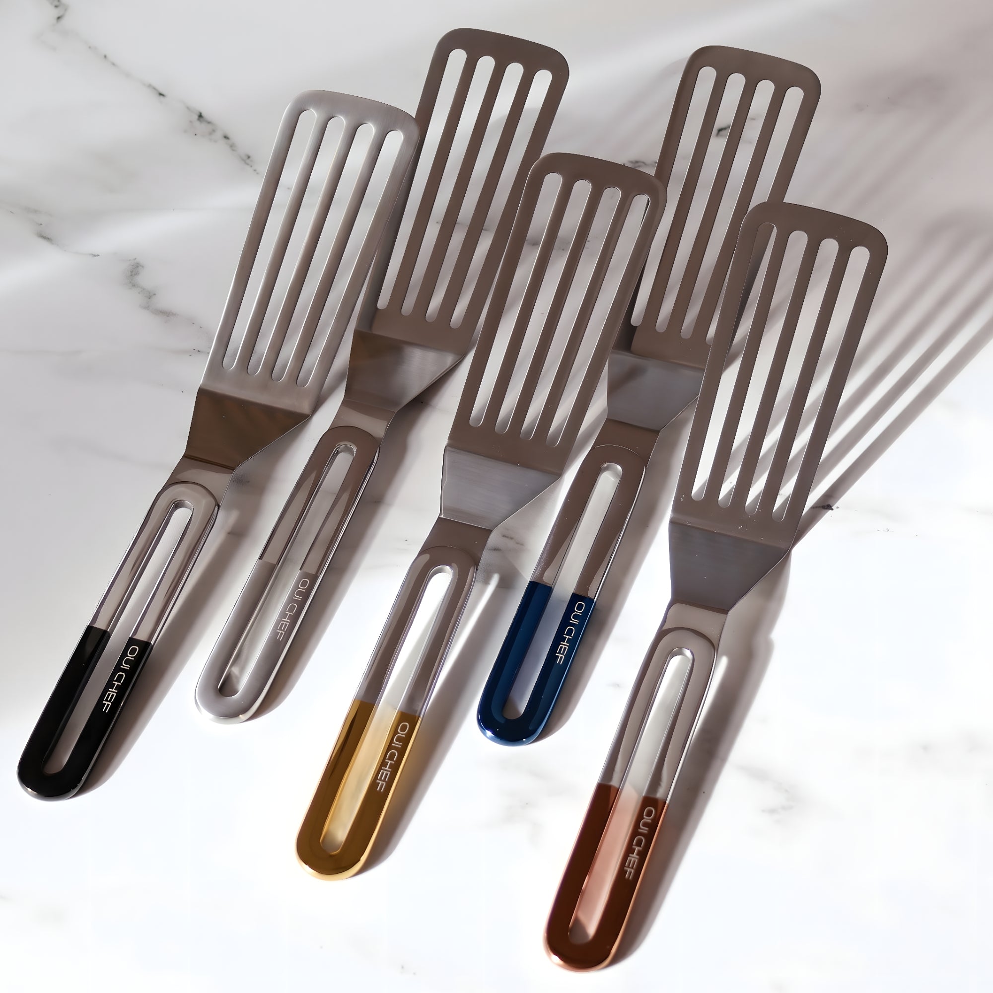 Five stainless steel spatulas with gloss black, gold, chrome, copper and metallic blue top handles arranged together on an elegant marble countertop in soft natural lighting, showcasing a premium modern kitchen aesthetic.