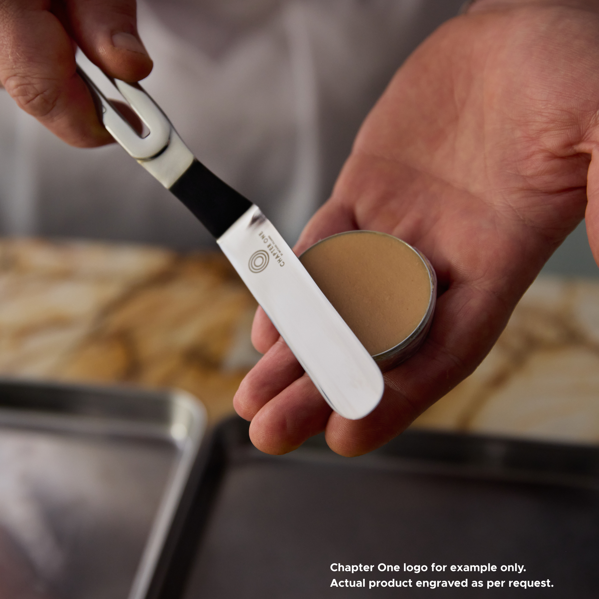 A close up of a Chef smoothing a tin of pate with a stainless steel palette knife that has an edge-to-edge sharp blade and the "Chapter One" logo engraved on it.