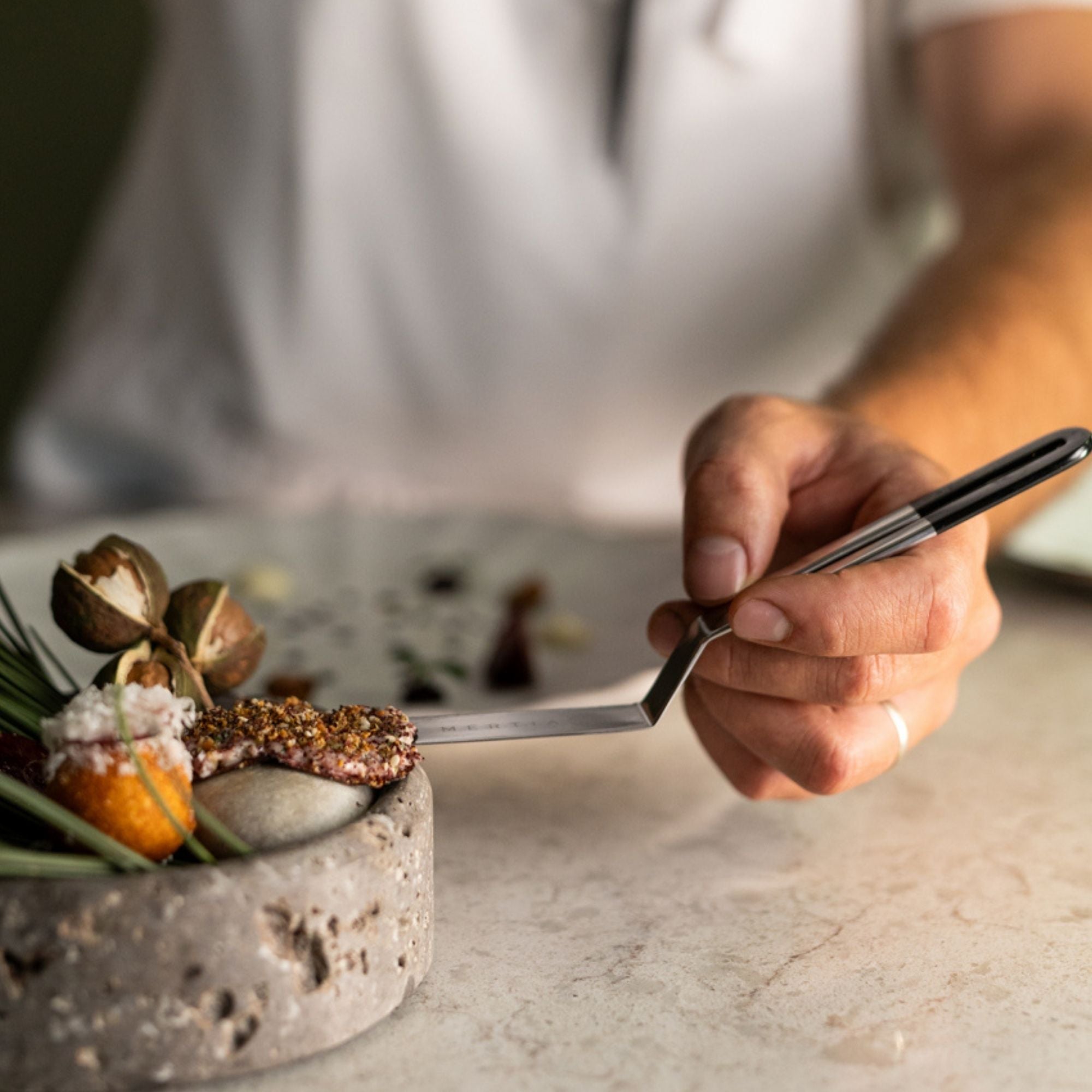 A Chef holding a stainless steel palette knife with a gloss black top handle in hand, plating a dish with garnishes on a marble worktop surface