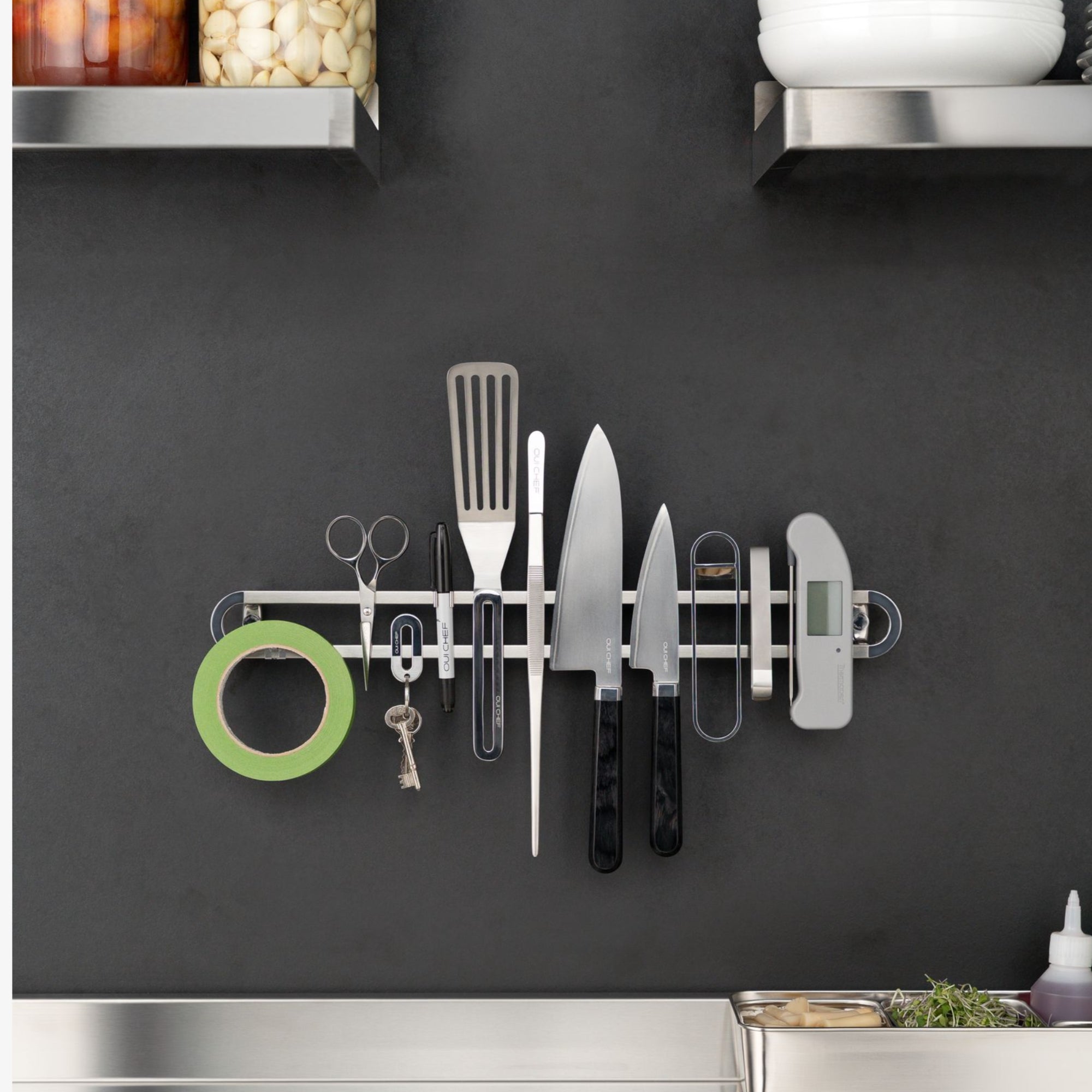 A Stainless steel magnetic placed on a charcoal coloured restaurant kitchen wall with the following line up from left to right; kitchen tape, scissors, keys, a sharpie, a spatula, tweezers, two chef's knives, a bottle opener, fish tweezers and a thermometer.