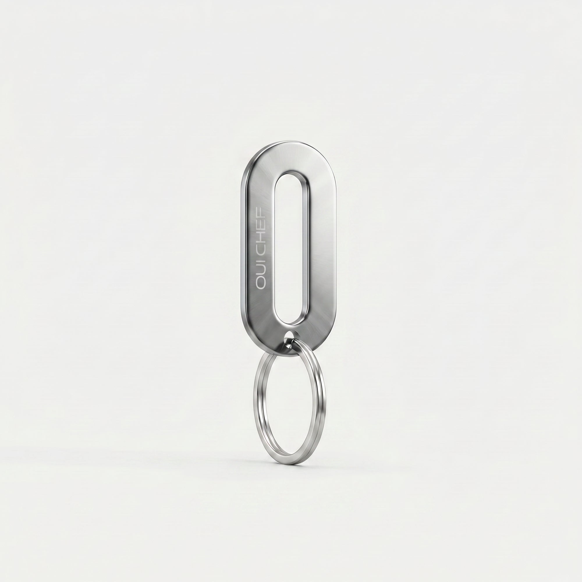 A chrome keyring engraved with the Oui Chef logo styled standing upright on its ring in an artistic pose, set against a soft off-white background with subtle shadows highlighting its glossy finish.