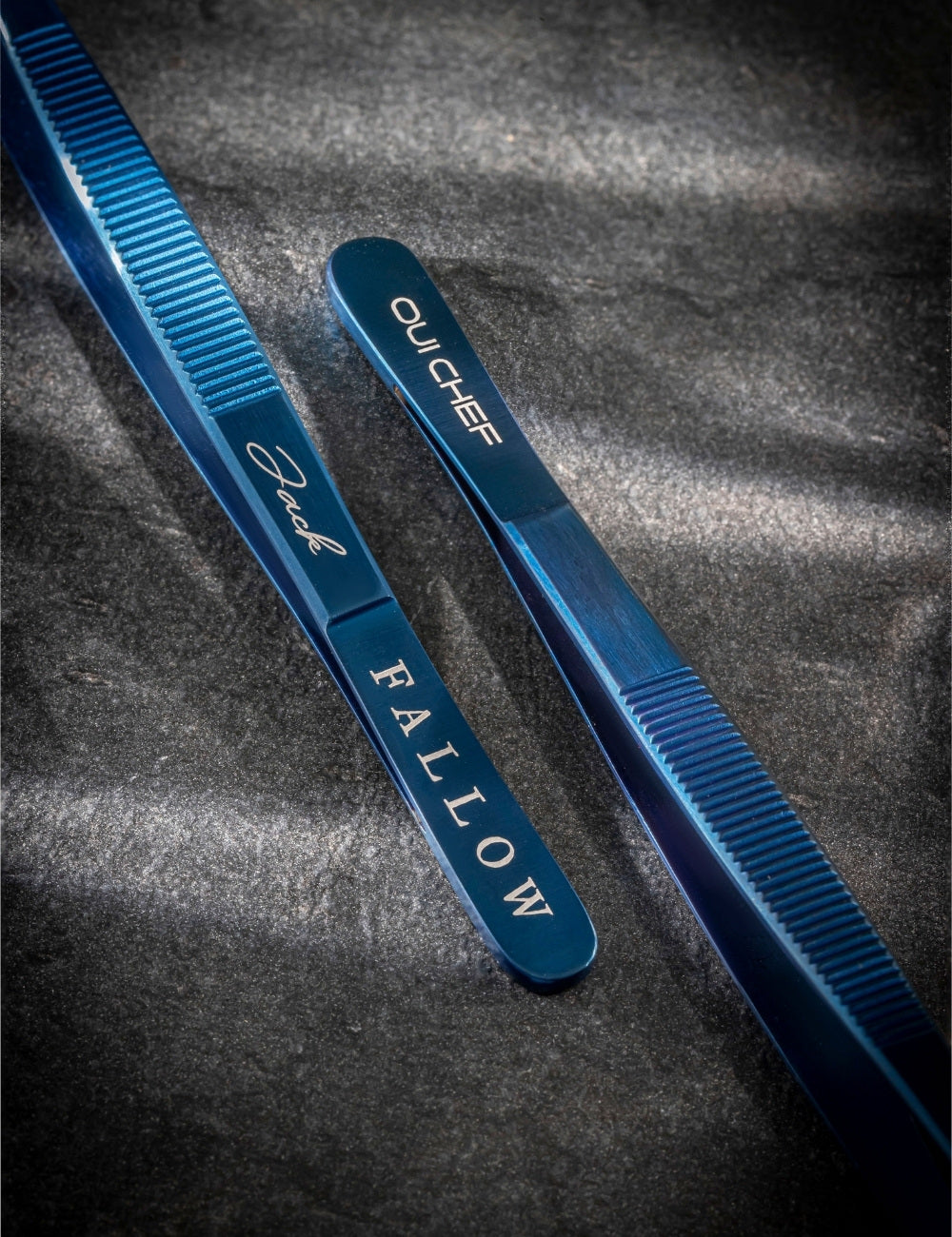 Metallic blue tweezers with 'Oui Chef' branding on one side and "Fallow" restaurant logo engraved on the other side placed on a textured dark background.