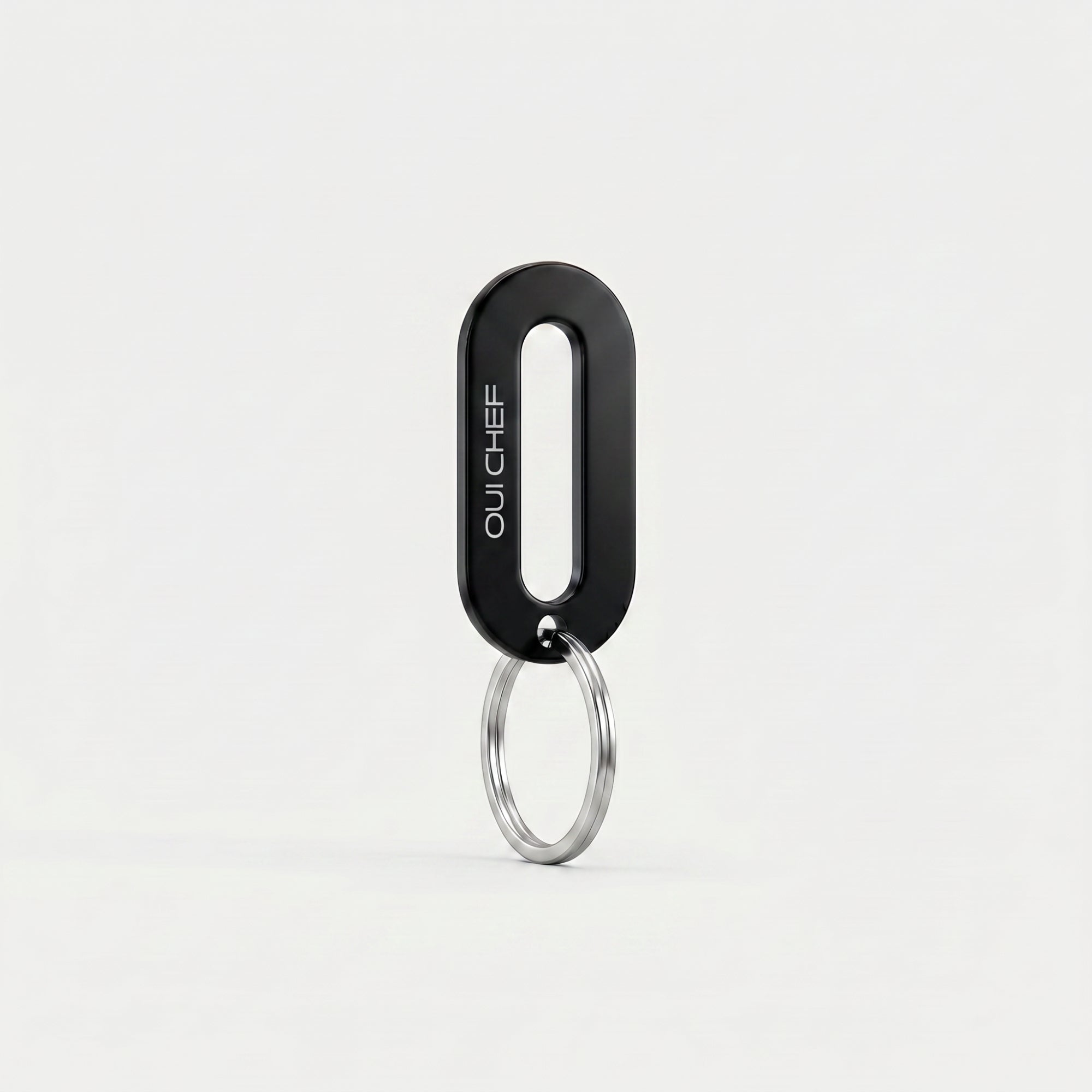 A gloss black keyring engraved with the Oui Chef logo styled standing upright on its ring in an artistic pose, set against a soft off-white background with subtle shadows highlighting its glossy finish.