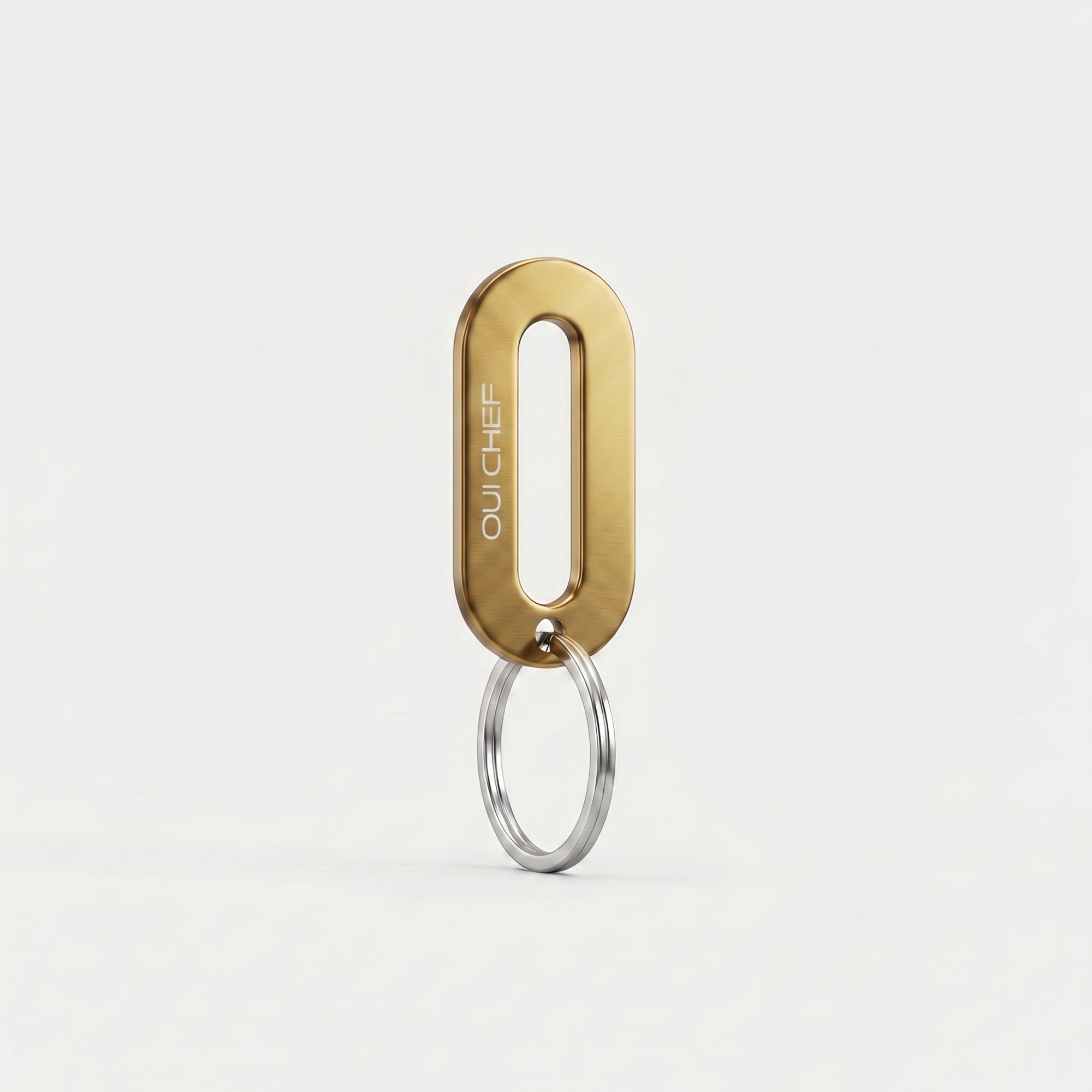 A gold colour keyring engraved with the Oui Chef logo styled standing upright on its ring in an artistic pose, set against a soft off-white background with subtle shadows highlighting its glossy finish.