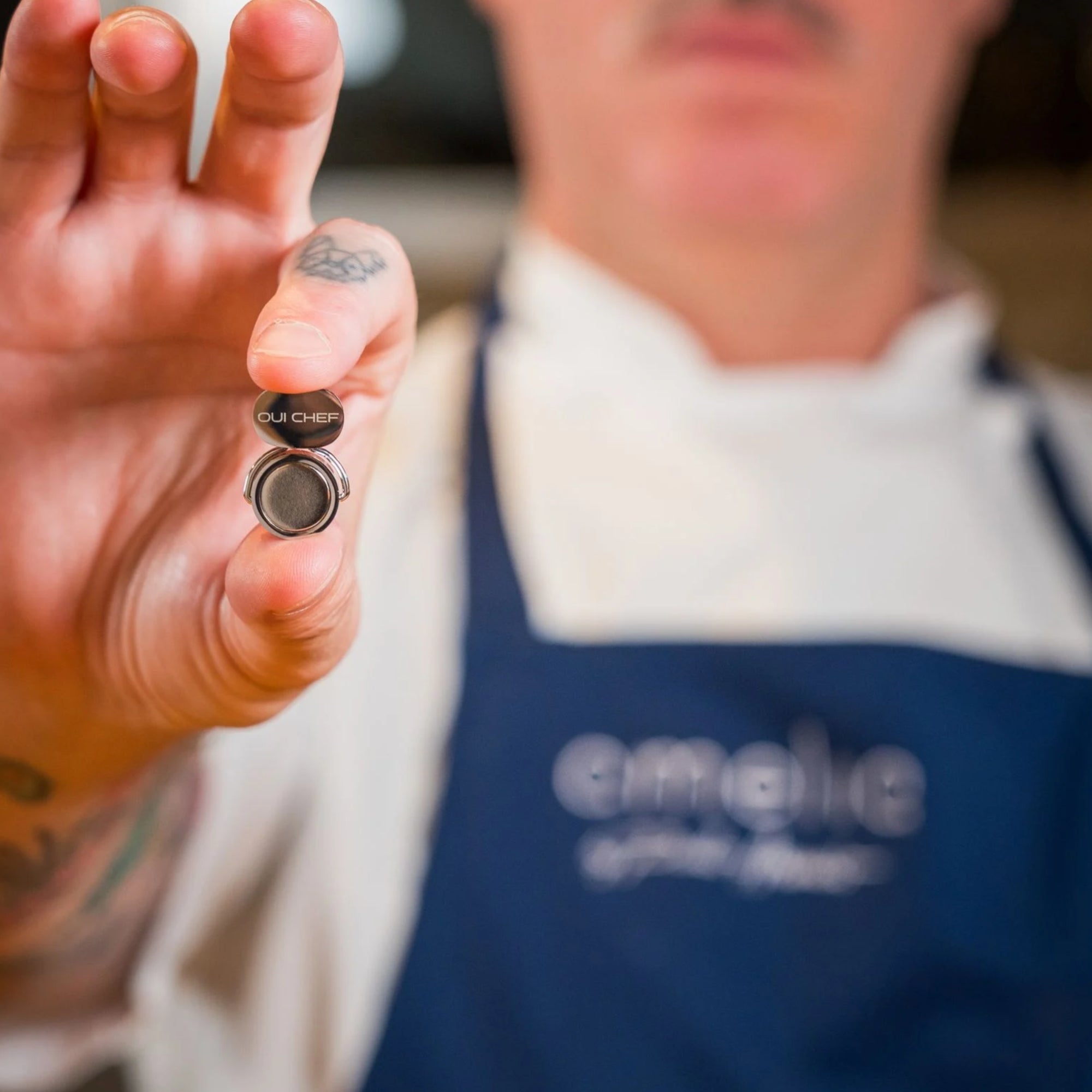 Chef holding a holdfast magnetic clip with 'OUI CHEF' branding, wearing a blue apron.