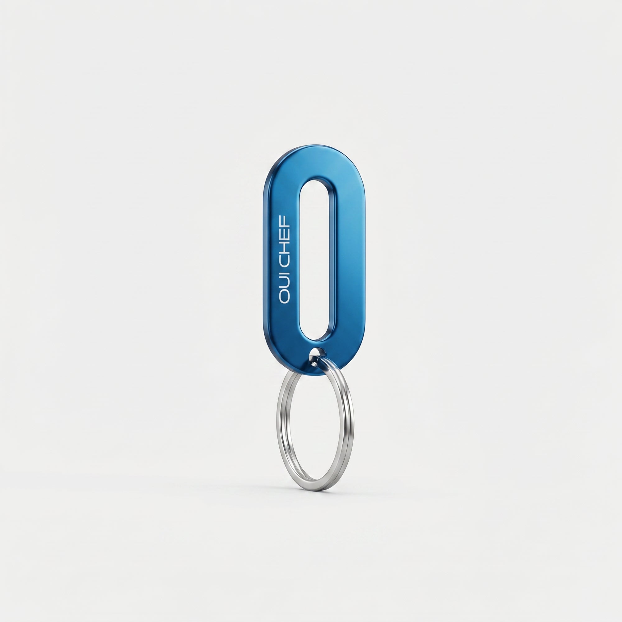 A metallic blue keyring engraved with the Oui Chef logo styled standing upright on its ring in an artistic pose, set against a soft off-white background with subtle shadows highlighting its glossy finish.