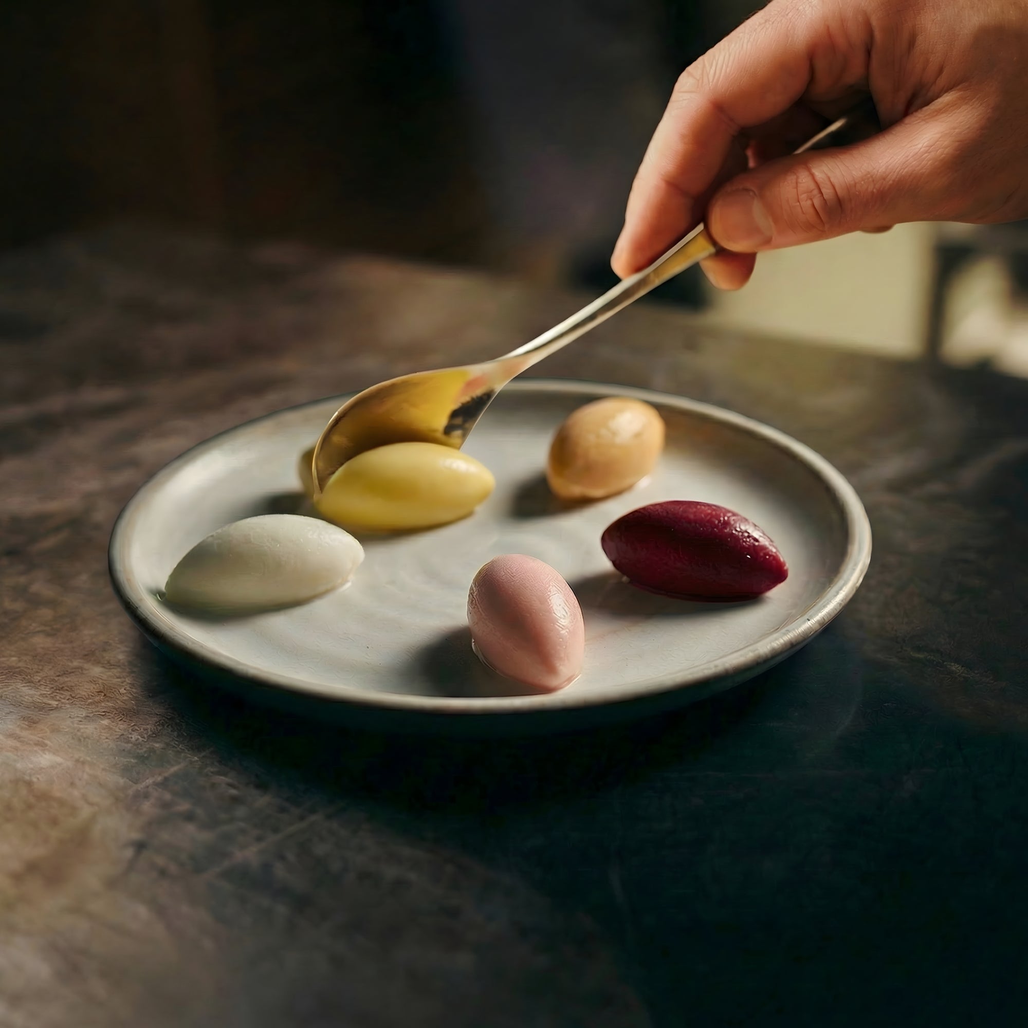 A chef carefully plating five differently coloured ice cream rochers with a small stainless steel rocher spoon onto a ceramic plate.