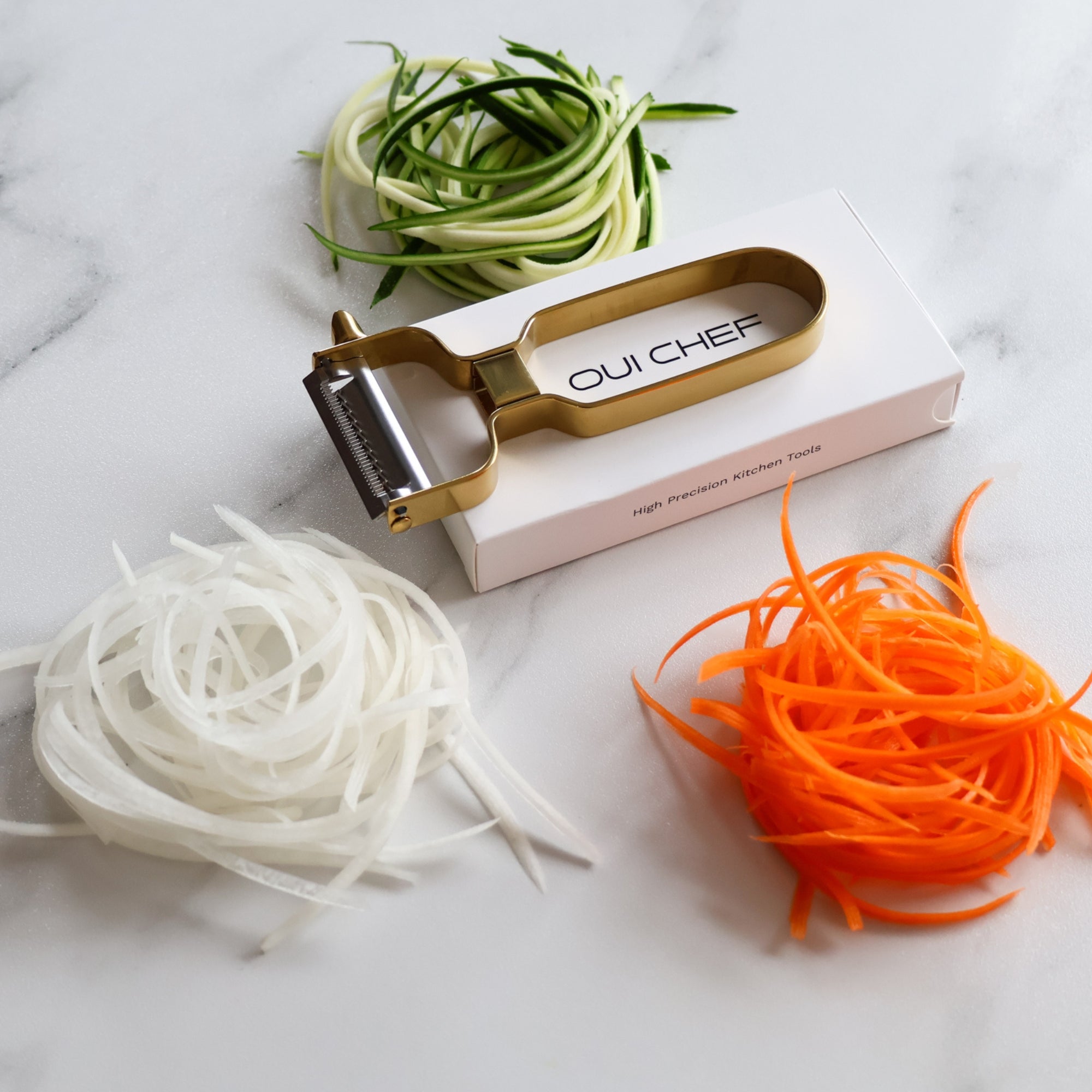 Oui Chef julienne peeler with nests of zucchini, carrot, and radish on a marble countertop.